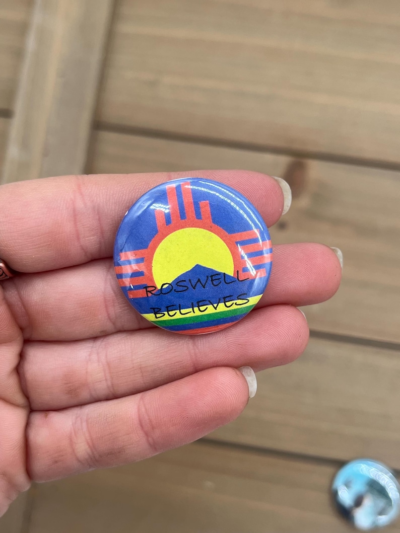 Peut inclure: Un bouton rond avec un fond bleu et un soleil jaune au centre. Le soleil est entour&eacute; d'un symbole rouge et orange. Le texte "Roswell Believes" est &eacute;crit en noir sous le symbole.