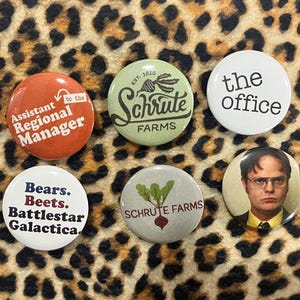 May include: A collection of six round buttons with various designs and text. The buttons feature phrases like "Assistant Regional Manager," "Schrute Farms," and "The Office." One button displays a portrait of a man. The background is a leopard print.