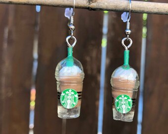 Peut inclure: Une paire de boucles d'oreilles argentées avec un charme en plastique transparent en forme de gobelet Starbucks Frappuccino avec un liquide brun et de la crème fouettée blanche. Le charme a un couvercle vert et un logo Starbucks vert avec une sirène blanche.
