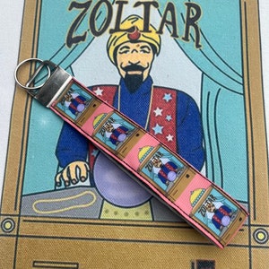 May include: A pink and white keychain with a repeating image of Zoltar, a fortune teller machine, with a silver key ring.