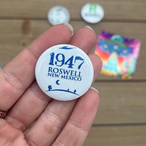 Peut inclure: Bouton blanc avec du texte bleu qui dit "1947 Roswell Nouveau-Mexique" avec un croissant de lune et des silhouettes de cactus.