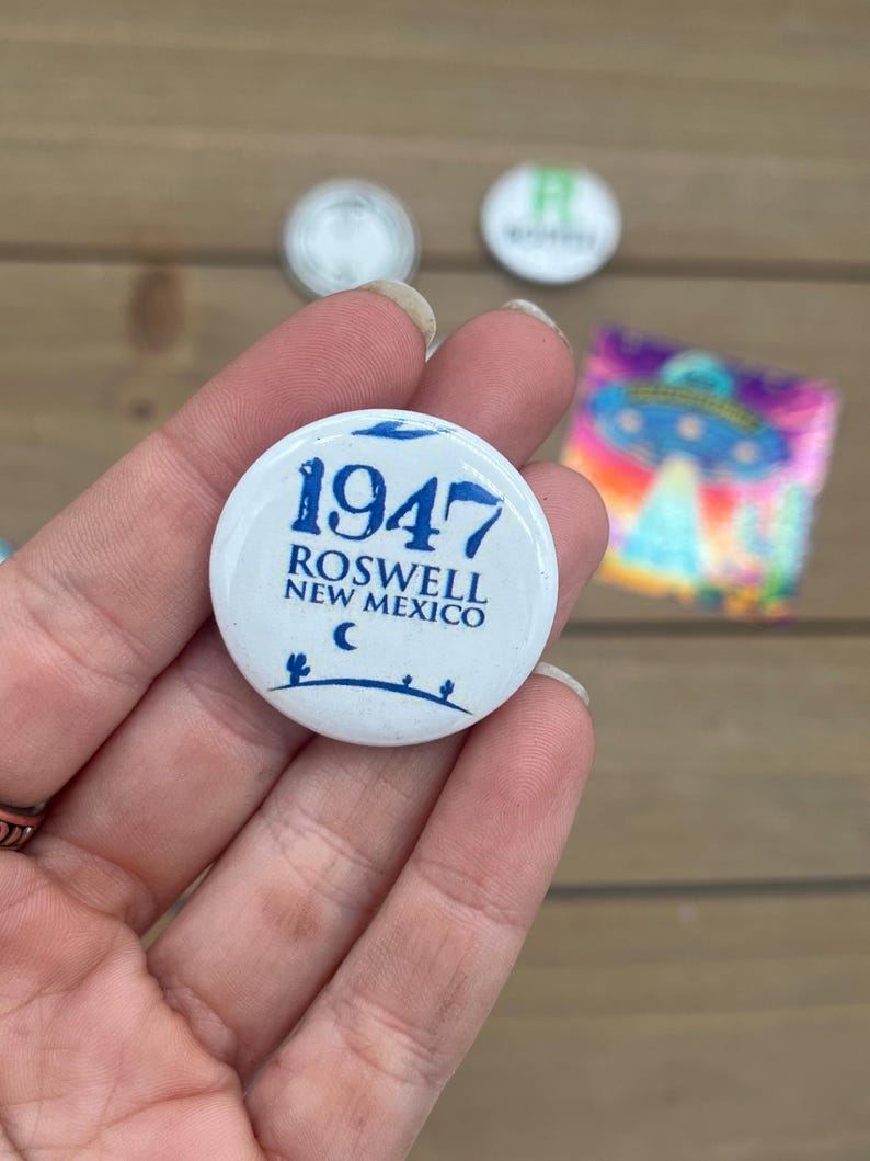 Peut inclure: Bouton blanc avec du texte bleu qui dit "1947 Roswell Nouveau-Mexique" avec un croissant de lune et des silhouettes de cactus.