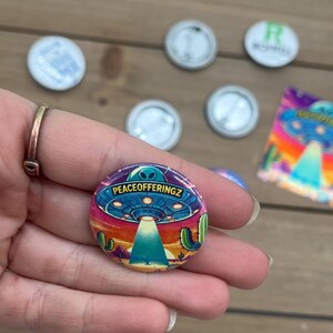 Peut inclure: Un bouton color&eacute; avec un OVNI de dessin anim&eacute; volant au-dessus d'un paysage d&eacute;sertique. Le texte "PEACEOFFERINGZ" est imprim&eacute; sur l'OVNI.