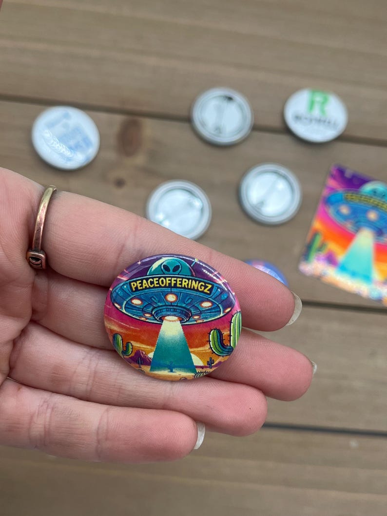 Peut inclure: Un bouton color&eacute; avec un OVNI de dessin anim&eacute; volant au-dessus d'un paysage d&eacute;sertique. Le texte "PEACEOFFERINGZ" est imprim&eacute; sur l'OVNI.