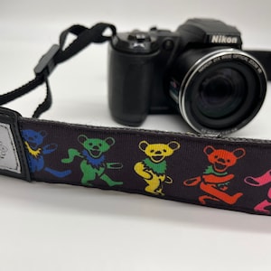 May include: A black camera with a zoom lens and a black camera strap with a colorful dancing bear pattern. The strap has the text "00200" on it.