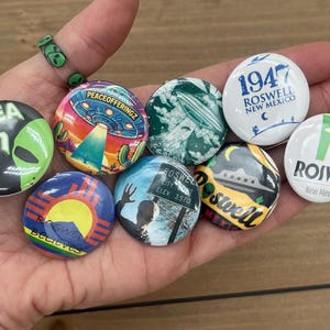 May include: A collection of nine colorful button pins featuring various designs related to Roswell, New Mexico, including the words "Area 51", "Roswell", "1947", and "Peace Offeringz".