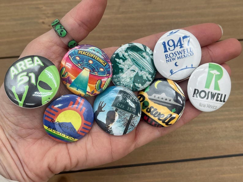 Peut inclure: Une collection de neuf badges boutonn&eacute;s color&eacute;s pr&eacute;sentant divers motifs li&eacute;s &agrave; Roswell, au Nouveau-Mexique, notamment les mots "Area 51", "Roswell", "1947" et "Peace Offeringz".