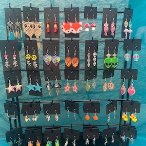May include: A black display rack showcasing a variety of earrings. The earrings feature diverse designs, including stars, hearts, mushrooms, aliens, and peace signs. Colors include pink, blue, green, and orange. The earrings are displayed on black cards.