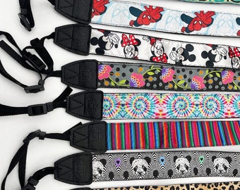 May include: Eight different camera straps with various colorful patterns. The straps are made of fabric and have adjustable black straps with a camera icon.