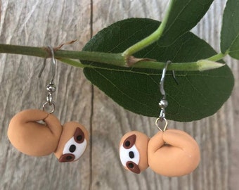 Peut inclure: Une paire de boucles d'oreilles pendantes représentant un paresseux brun avec des yeux blancs et des accents bruns suspendus à des crochets argentés.