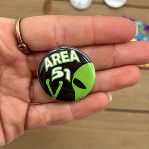 Peut inclure: Un bouton noir et vert avec le texte "AREA 51" et un visage d'extraterrestre vert.