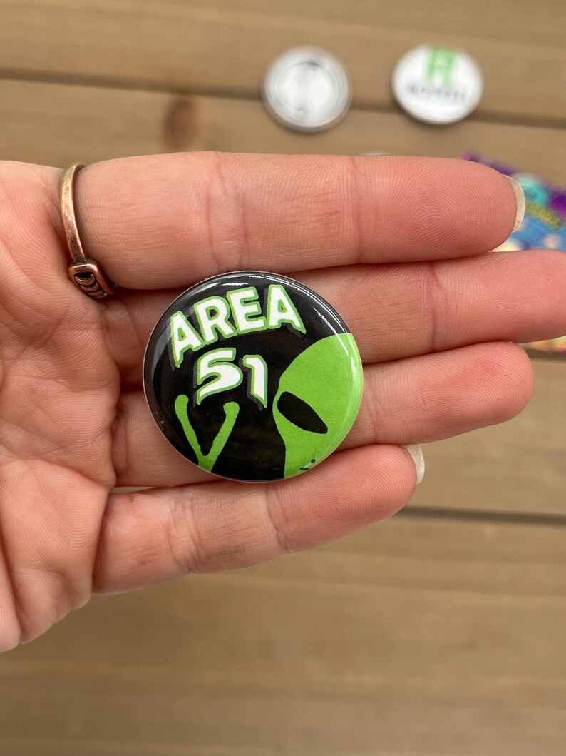 Peut inclure: Un bouton noir et vert avec le texte "AREA 51" et un visage d'extraterrestre vert.