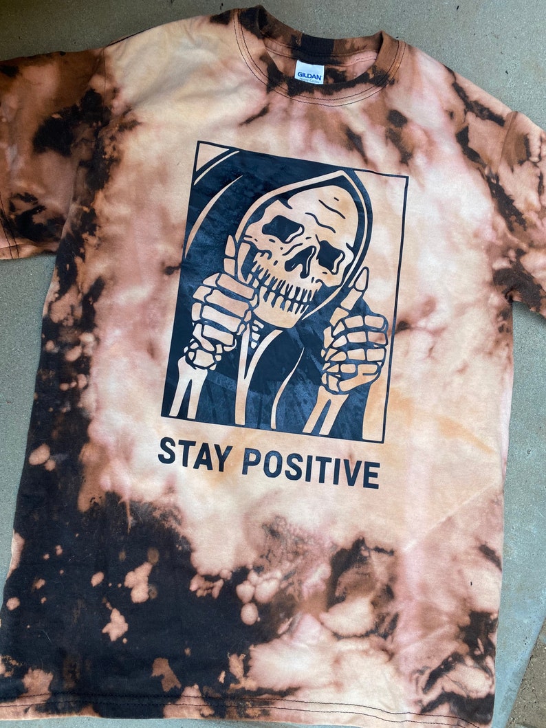 May include: A black and white tie-dye t-shirt with a graphic of a skeleton giving a thumbs up. The graphic is in a black rectangle with the text "STAY POSITIVE" below it.