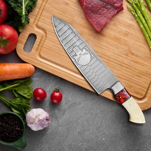 K&ouml;nnte beinhalten: Ein Edelstahl-Kochmesser mit einem Sch&auml;del-Design auf der Klinge und einem rot-wei&szlig;en Griff. Das Messer liegt auf einem Holzbrett mit einem St&uuml;ck rohem Fleisch und Spargel.