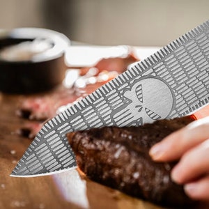 K&ouml;nnte beinhalten: Ein silbernes Messer mit einem in die Klinge ge&auml;tzten Sch&auml;delmuster. Das Messer wird zum Schneiden eines Fleischst&uuml;cks verwendet.