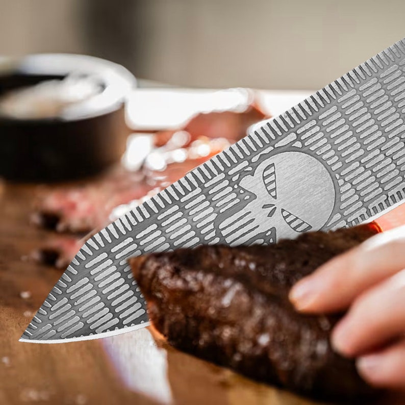K&ouml;nnte beinhalten: Ein silbernes Messer mit einem in die Klinge ge&auml;tzten Sch&auml;delmuster. Das Messer wird zum Schneiden eines Fleischst&uuml;cks verwendet.