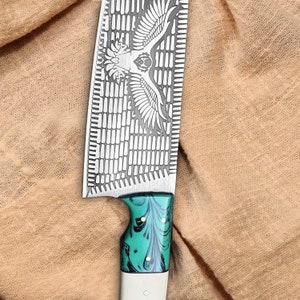 K&ouml;nnte beinhalten: Ein gro&szlig;es Kochmesser mit einer gemusterten Klinge, die ein Adlerdesign zeigt. Der Griff ist aus wei&szlig;em und gr&uuml;nem Holz mit einem wirbelnden Muster.