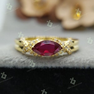 Könnte beinhalten: Ein goldener Ring mit einem roten, markisenförmigen Edelstein, der von kleineren weißen Edelsteinen umgeben ist. Der Ring hat ein zartes, filigranes Design.
