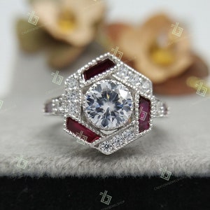 Puede incluir: Un anillo de plata con una piedra central grande, redonda y transparente rodeada de piedras transparentes más pequeñas y acentos rojos. El anillo tiene una forma hexagonal.