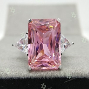 Könnte beinhalten: Ein Verlobungsring aus Silber mit einem pinkfarbenen Smaragdschliff und zwei kleineren Diamanten als Akzente auf beiden Seiten.