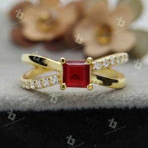 Peut inclure: Une bague en or avec une pierre carrée rouge et de petites pierres blanches sur la bande.