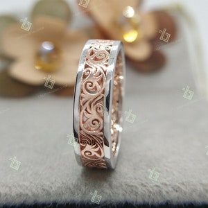 May include: A close-up of a wedding band with a rose gold band and a white gold band. The rose gold band has a decorative filigree design.