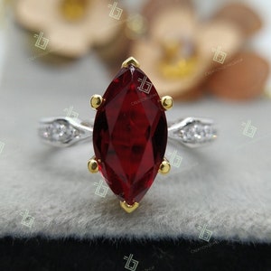 May include: A marquise-shaped red gemstone ring set in a silver band with a gold accent. The ring features small clear gemstones on the band.