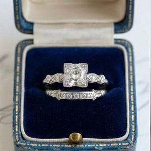 Puede incluir: Un conjunto de anillos de compromiso en tono plateado con una piedra central de corte cuadrado flanqueada por piedras redondas más pequeñas en una banda decorativa, junto a una alianza a juego con cinco piedras redondas, en un estuche de terciopelo azul.