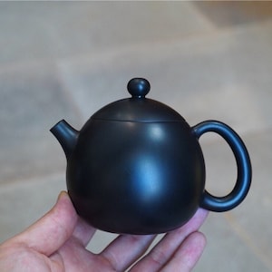 May include: A black ceramic teapot with a rounded body and a small knob on the lid. The teapot has a spout on one side and a handle on the other.
