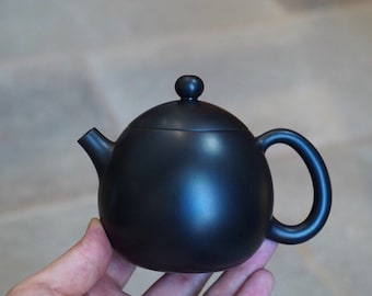 May include: A black ceramic teapot with a rounded body and a small knob on the lid. The teapot has a spout on one side and a handle on the other.