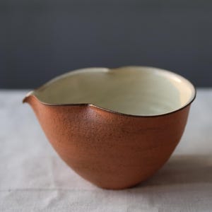 May include: A handmade, terracotta-colored ceramic bowl with a unique, asymmetrical shape. The interior is a creamy off-white, contrasting with the textured exterior. The bowl features a small spout and a dark rim.