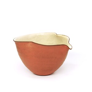 May include: A terracotta-coloured ceramic bowl with a cream-coloured interior and a unique spout. The bowl has a rounded shape and a textured surface. The rim is outlined with a dark brown colour. The bowl is set against a white background.