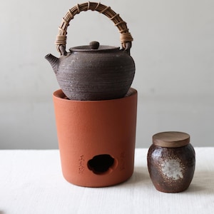 May include: A dark brown textured ceramic teapot with a woven rattan handle sits atop a cylindrical terracotta stove featuring a cutout opening and embossed Chinese characters. Beside it is a small, dark brown glazed jar with a flat wooden lid.