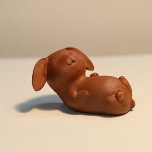 Könnte beinhalten: Kleine, braune Keramikfigur eines Hundes, der auf dem Rücken liegt und die Pfoten in die Luft reckt. Der Hund hat Schlappohren und einen lächelnden Gesichtsausdruck. Die Figur ist einfarbig und scheint handgefertigt zu sein.
