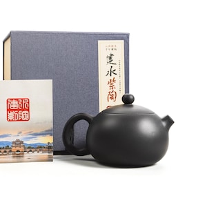 Könnte beinhalten: Eine schwarze Teekanne aus Keramik mit Deckel und Ausguss. Die Teekanne steht auf einer weißen Oberfläche. Die Teekanne steht vor einer blauen Box mit chinesischen Schriftzeichen. Die Box ist mit "Wasserkocher" beschriftet.