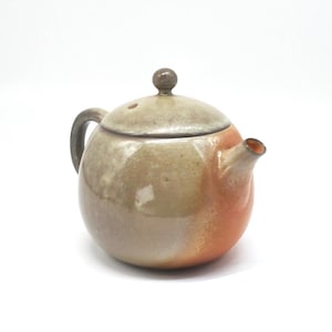 May include: A ceramic teapot with a rounded body and a lid. The teapot is primarily tan with orange accents near the spout. It has a dark brown handle and a small knob on the lid. The teapot is designed for brewing tea.