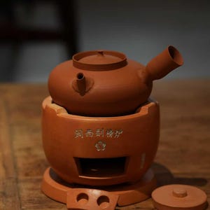 以下が含まれることがあります： テラコッタ色のティーセット。注ぎ口と蓋が付いたティーポットと、小さな開口部と別の蓋が付いたお揃いのベースが含まれています。木製の表面にセットが置かれています。