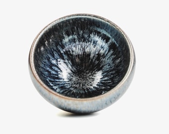 Puede incluir: Una taza de té pequeña de cerámica azul oscuro con un esmalte moteado blanco y negro. La taza es redonda y tiene un cuenco ancho y poco profundo.