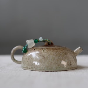 May include: A miniature ceramic teapot with a speckled, beige glaze. It features a curved handle and a short spout. The lid is decorated with a beaded accent of green and white beads. The teapot is set against a neutral background.