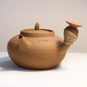 May include: A brown ceramic teapot with a rounded body, a spout, and a lid with a small knob. The handle is wrapped with twine. The teapot has a small inscription on the side. The teapot is likely used for brewing tea.