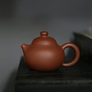 Può includere: Una piccola teiera in ceramica marrone con un corpo arrotondato, un manico curvo e un piccolo beccuccio. Il coperchio ha un pomello arrotondato. La teiera è su una superficie scura.