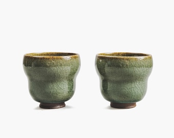 May include: Two green ceramic teacups with a crackled glaze finish. The cups have a rounded, slightly flared shape.