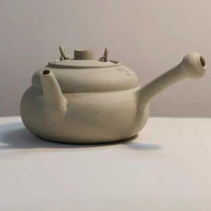 Peut inclure: Une théière en céramique grise claire faite à la main, avec un corps arrondi, un long bec incurvé et un couvercle avec deux petites poignées. La théière a un design simple et rustique.