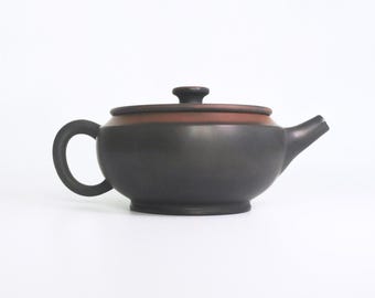 May include: A dark grey ceramic teapot with a brown rim on the lid and a curved handle. The teapot has a spout and a small knob on the lid. The teapot is on a white background.