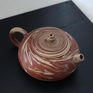 May include: A round, ceramic teapot with a swirl pattern in shades of brown and cream. The teapot has a handle, a spout, and a small lid. The design features a spiral pattern across the top.