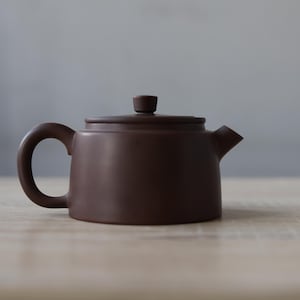 May include: A small, brown ceramic teapot with a rounded body, a curved handle, and a spout. The lid has a small knob. The teapot is on a light-coloured wooden surface. The background is a soft grey.