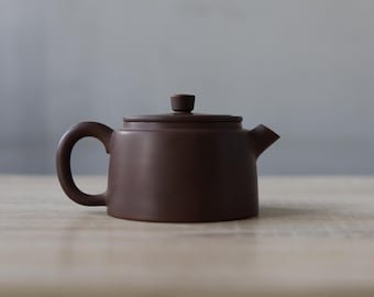 May include: A small, brown ceramic teapot with a rounded body, a curved handle, and a spout. The lid has a small knob. The teapot is on a light-coloured wooden surface. The background is a soft grey.