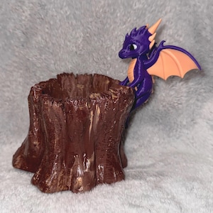 Puede incluir: Una maceta marrón en forma de tocón de árbol con una figura de dragón púrpura posada en el lateral. El dragón tiene alas y cuernos naranjas. La maceta está hecha de un material marrón oscuro con una superficie texturizada.