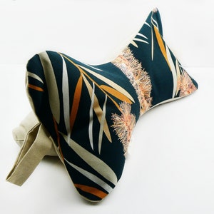 May include: A decorative lumbar pillow with a botanical print. The pillow features a dark teal background with a pattern of orange, beige, and white leaves and floral accents. A beige fabric handle is attached to the pillow.