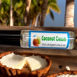 May include: A clear glass roll-on bottle with a black cap and a blue label that says "Coconut Cream" and "TheCrabAppleCabin.com". The bottle is sitting on a wooden surface next to two coconut cream pies.